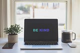 A desk with a laptop in the middle with the words “BE KIND” on the screen.