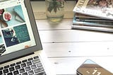 A workdesk with a MacBook, mobile phone, books and a vase.