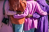 Four women from behind with arms around each other’s backs, wearing colorful pink, purple and orange sweaters!