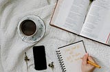 A coffee mug, phone, journal, and Bible sit on a white blanket.