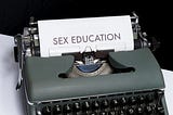 A grey typewriter with paper titled sex education sits on a white desk.