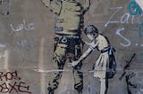 Graffito on a wall, showing a soldier with his back turned towards the viewer and his hands in the air. A little girl is performing a body search on the soldier, making the picture a subversion of usual expectations.