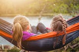 Two young girls sitting on a hammock with their backs to us.
