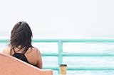 A woman from behind, who has dark hair down to her shoulders, and is wearing a black halterneck vest top. She is sitting in a pink seat with a takeaway coffee next to her and she is looking out at the sea. There is an aqua-blue divide between her and the sea, like she is on a boat.