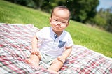 Asian baby sitting on a plaid blanket over grass. He is ostensibly frowning.