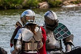 Two people in full plate armour standing by water.