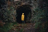 A woman in a strong yellow raincoat standing at the entrance of a large cave looking at its darkness. We all go though dark moments. But can come out strengthened.