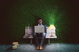 A man is sitting on a bench in a park, typing on a laptop. There’s a coffee and a lit lamp next to him.