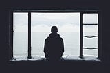 A silhoutte of a person sitting on a snow-covered windowsill of an abandoned building, looking out over a body of water on an cloudy day.