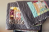 A picture of a folded quilt. It appears to be laying over the back of a chair, and the colors in the quilt include grey, blue, orange, red, maroon, gold/yellow and turquoise.