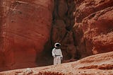 An astronaut in a white space suit walks through a desolate red landscape, dwarfed by the rocks behind them.