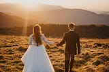 A groom leads a bride by the hand across a field at sunset.
