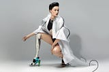 Cyborg Artist Viktoria Modesta crouching down showing off her metal leg in a heel, wearing a long white trench coat