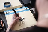 An illustrator tightly grips an ink pen as they draw a cartoon on a sheet of high quality expensive paper, which is held to a board with masking tape. His tools and materials and scattered around.