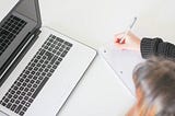 Adjacent to an open laptop computer, there is a notebook. The top of a person’s head is visible as well as their arm preparing to write on the notebook.