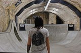 Image of a man with long hair, back to the camera, staring out over an indoor skate park.