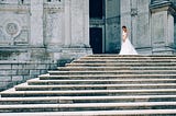 A woman in a wedding dress standing alone at the top of stairs at a church. Her head is down and her body language says she is disappointed and sad. This signifies the unmet expectations we can experience during separation on the Twin Flame journey.