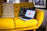 A colour photo of an open laptop on a yellow couch with a blanket and a cushion.