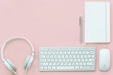 White keyboard, headphone, mouse and a copybook on a pink background, the photo have a modern calm feeling