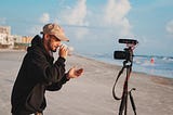 a guy on a beach standing in front of a camera looking into the camera with his hands towards his face