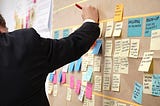 Man in a suit writes something down on a board with a bunch of sticky notes