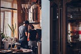 A couple stand in a kitchen in front of a stove cooking together with backs to the camera