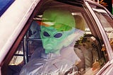 Person in driver seat of a car wearing an alien costume