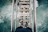 The view of the picture is a person looking down at his feet over a wooden bridge with a river underneath him.