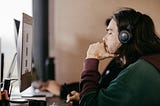 A man with long hair is sitting in front of a computer screen. He is wearing a headset, and his expression conveys deep thinking.