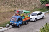 Looking down at a road with two parked vehicles. One is an ordinary white car. The other is a small blue cabbed truck with a traditional Chinese structure on top, with loudhailers either side pointing front and back. Behind the vehicles, a dry, scrubby grass bank slopes upwards