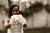 Lovely, smiling brown woman in a wedding dress with a bouquet of white and pink flowers in her out-stretched hands.