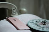 Corner of white bed with pink iphone 8 and alarm clock face lay across it