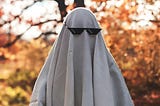 A creepy person standing in a park and covered with a white sheet to look like a ghost wearing sunglasses.