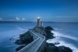 A lighthouse at the end of a long walkway begins to shine at dusk.