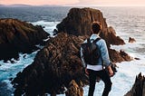 A man with a backpack stands looking out over a breathtaking view of ocean cliffs.