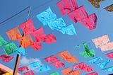 Against a bright blue sky, and colorful pieces of cut paper are hung along wires suspended in the air. Red, orange, blue, green and white, these decorative paper pieces flap in the wind like flags