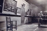 Black and white photo of an art gallery with classic paintings in elaborate frames, a simple wooden chair and a man sitting thoughtfully in the corner.