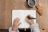 A woman writing in a journal, while eating a croissant and drinking coffee