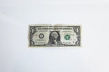 A photo of a US 1-Dollar bill sitting face-up on a flat, white surface.