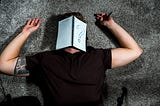 A guy laying down on a carpeted floor with a notebook on his face with a question mark.