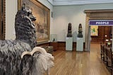 Statues of an alpaca and a goat look into an art gallery with large oil paitnings, statues on plinths, a wooden bench. At the far end of the gallery a large classical doorway has a sign above it that reads ‘people’