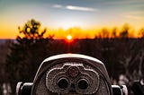 In-focus coin operated binoculars overlooking a blurry backdrop of the sunset and large trees.