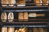 Dark-stained, open wooden shelves at a bakery display at least nine different varieties of bread. Visible handwritten chalk strip signage indicates  Spelt Sourdough, Honey Spelt Bread, Simple Sourdough, 4-Grain Pan Bread, and Ancient Multigrain Pan Bread, with prices. Two of the other varieties look like baguettes and focaccia.