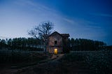 A haunting two-story farmhouse in a desolate wood. The front door is open, light on in an otherwise dark farmhouse.