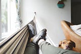 A pair of jogger-clad legs in sneakers propped up in a hammock with a brown dog peering at the camera.