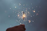 A dark hand holding up a short sparkler nearing the end of its sparks. Yellow sparks are seen flying across the purely medium grey background.