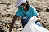 A masked person in a white hat is collecting trash off the beach. They are digging in the sand, filling a bag up with discarded cans and various pieces of garbage in an attempt to improve the natural landscape.