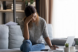 Woman, looking worriedly at a laptop and a notebook.