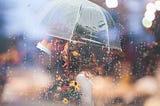 A man and a woman standing close to each other under umbrella because it is raining. The woman is whispering something in man’s ear.