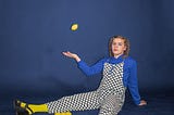 Lady in chequered dungarees over a blue sweater and yellow socks, sitting on the floor, juggling one lemon with others on the floor around her.